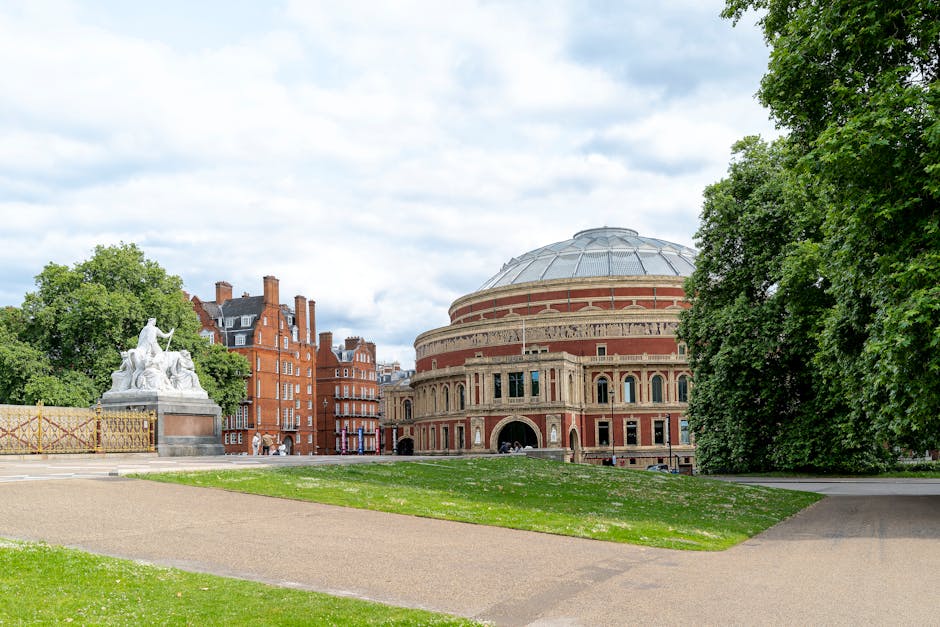 A wide view of the Royal Albert Hall, a large circular concert hall with a distinctive domed roof made of metal and glass panels. In the foreground, there is a well-maintained grassy area and a wide gravel pathway. To the left, a white sculpture depicting a seated figure alongside a winged lion, both made of stone or marble, is partially obscured by green foliage from nearby trees. The background shows historic red brick buildings with chimneys, and the scene is set under a partly cloudy sky with patches of blue. The area appears clean and well-kept, typical of the surroundings around Victoria and Albert Museum in South Kensington, London. South Kensington Cleaners specializes in surface cleaning and deep cleaning, ensuring hygiene and maintenance of such areas.