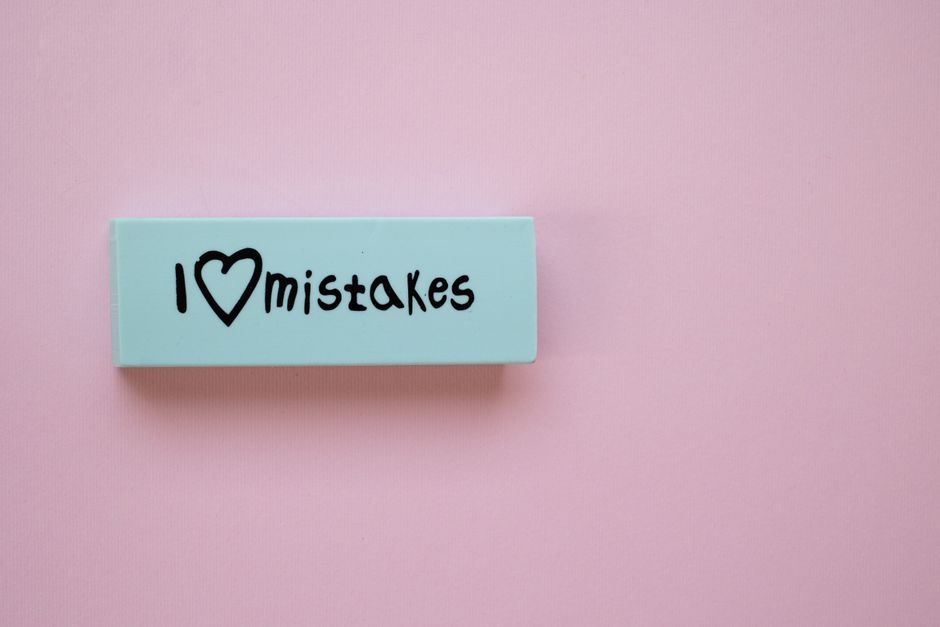A light blue and white eraser with the handwritten phrase 'I ❤️ Mistakes' is positioned on a soft pink background, alongside a white decorative pencil with small gray dots and a black tip. Below, 