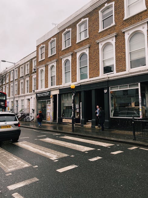 A street scene in South Kensington featuring a brick building with white window trims and large glass storefronts on the ground floor. The sidewalk in front is wet, indicating recent cleaning or rain,