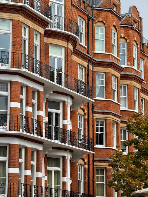Exterior view of a red brick residential building with multiple floors and white accents around the windows and columns, featuring black wrought iron balconies. The building's facade appears clean and well-maintained, with large windows reflecting the natural daylight. In front of the building, a tree with green and yellow leaves adds a touch of nature. The image emphasizes a tidy and presentable appearance typical of properties serviced by South Kensington Cleaners, known for their same-day cleaning offerings at South Kensington station SW7. The overall scene conveys an urban residential setting with a focus on building exterior cleanliness and maintenance, consistent with professional surface cleaning and upkeep services.