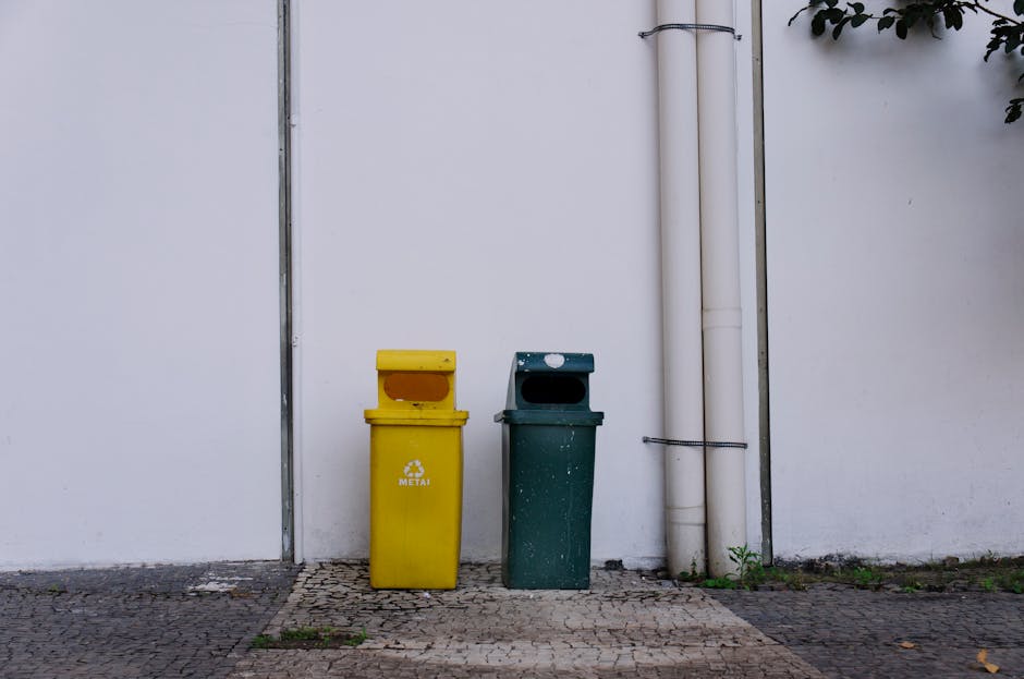 A person wearing a white shirt and transparent disposable gloves is holding a white plastic trash bin labeled 'TRASH' with black marker. The bin is lined with a black garbage bag, which is tied at the
